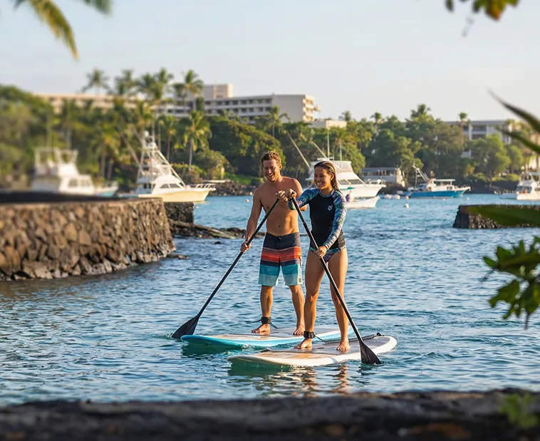 Two people paddleboarding in Keauhou Bay with boats and palm trees in the background
